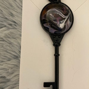 May include: A black metal key pendant with a round, black resin charm featuring a swirling abstract design in shades of purple, white, and black.