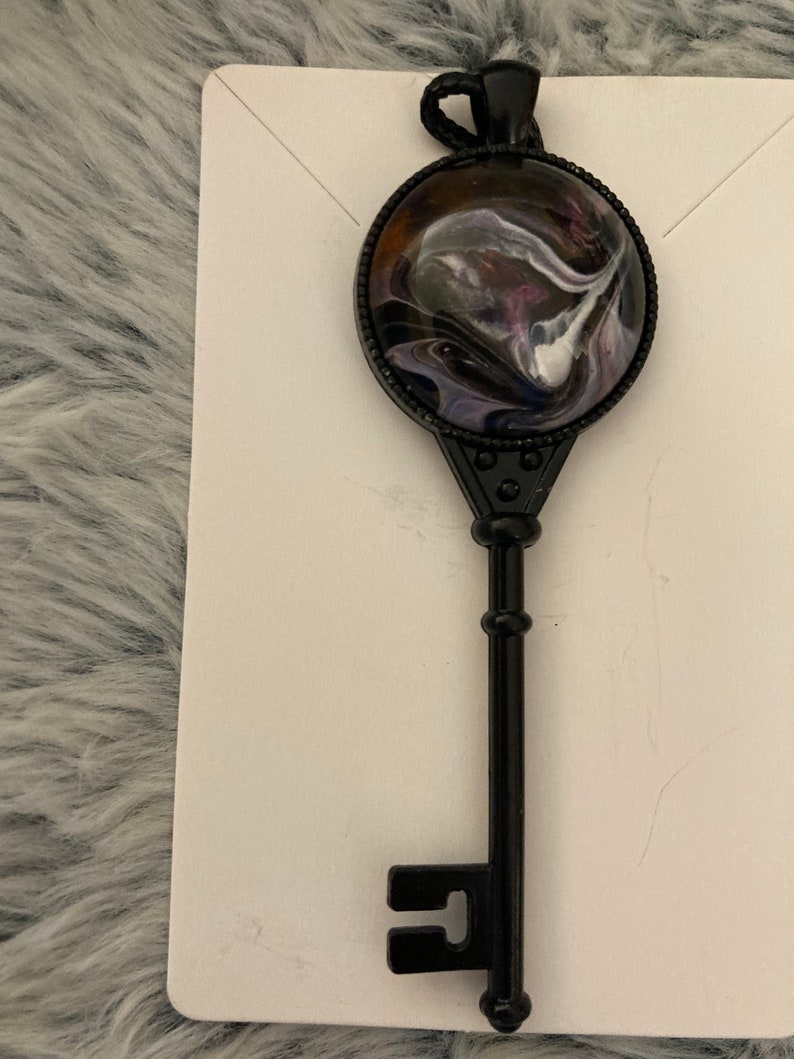 May include: A black metal key pendant with a round, black resin charm featuring a swirling abstract design in shades of purple, white, and black.