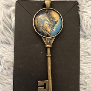 May include: A gold key pendant with a round blue, gold, and black resin inlay. The key is on a black display card.