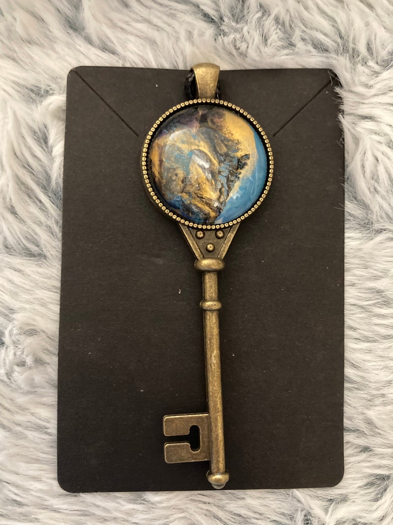 May include: A gold key pendant with a round blue, gold, and black resin inlay. The key is on a black display card.