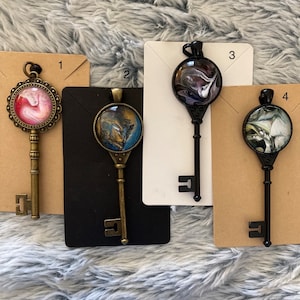 May include: Four key-shaped pendants with different abstract designs. Each pendant is made of metal and has a glass cabochon with a unique swirl pattern. The pendants are numbered 1 through 4.