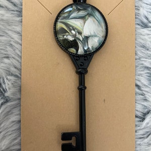 May include: A black metal key pendant with a round glass charm featuring a swirling black, white, and green marble design.