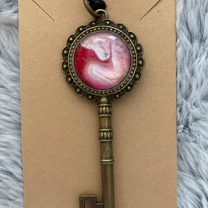 May include: A gold key pendant with a round, ornate frame. The frame encloses a pink and white abstract design with gold accents.