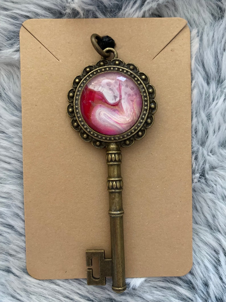 May include: A gold key pendant with a round, ornate frame. The frame encloses a pink and white abstract design with gold accents.