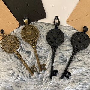 May include: Three decorative key pendants, two black and one gold, with intricate designs and heart accents. The keys are displayed on a fuzzy white surface.