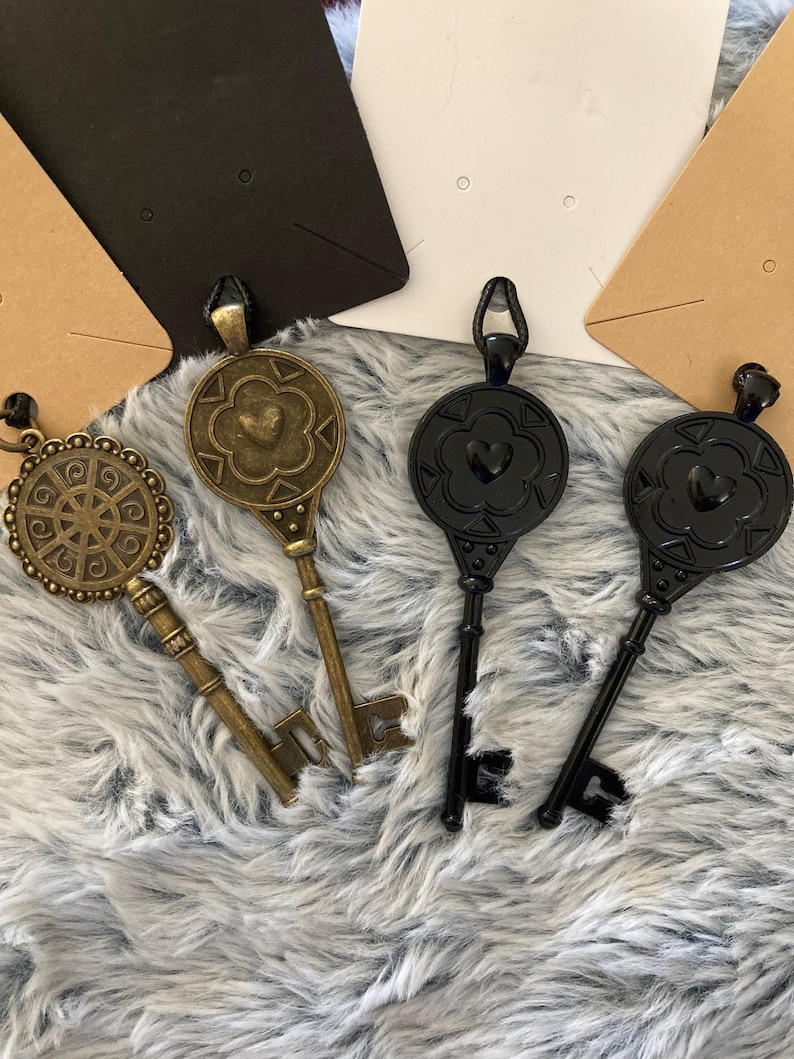 May include: Three decorative key pendants, two black and one gold, with intricate designs and heart accents. The keys are displayed on a fuzzy white surface.