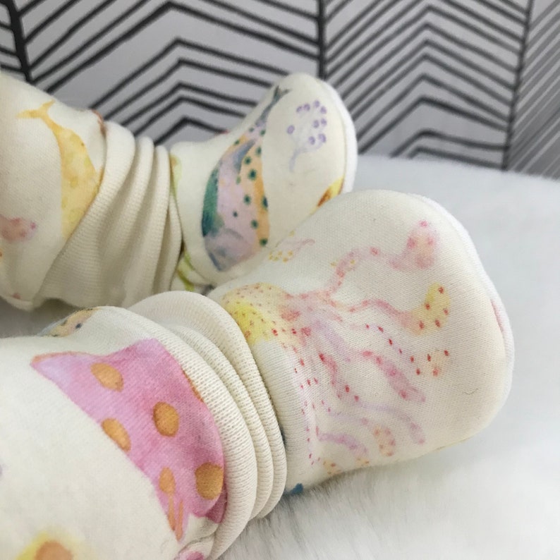 May include: A pair of white baby booties with a colorful sea creature print. The booties feature a yellow, pink, and blue watercolor design of fish, coral, and other sea life.