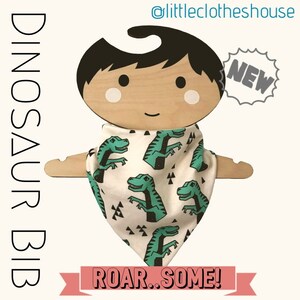 May include: A white bandana bib with a green dinosaur print and the text "Roar...Some!"  The bib is on a wooden doll hanger with the text "NEW" in a starburst shape.
