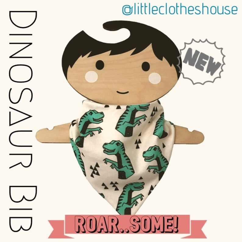 May include: A white bandana bib with a green dinosaur print and the text "Roar...Some!"  The bib is on a wooden doll hanger with the text "NEW" in a starburst shape.