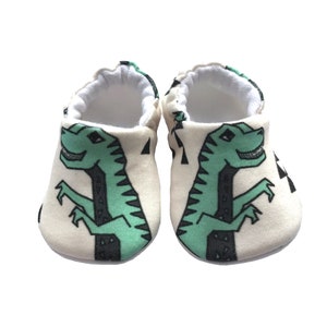 May include: A pair of white baby booties with a green dinosaur print. The dinosaurs are outlined in black and have a white background.