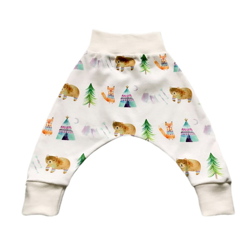 May include: White baby pants with a woodland animal print featuring bears, foxes, and teepees. The pants have a cream colored waistband and cuffs.