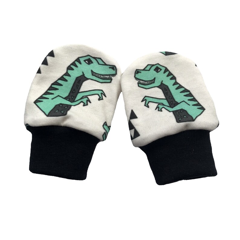 May include: A pair of white baby mittens with black cuffs. The mittens have a green dinosaur print with black outlines and white triangle patterns.