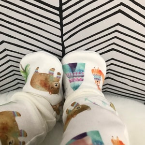 May include: White baby booties with a brown bear, a teepee, and a fox print. The booties are on a white surface with a black and white chevron pattern in the background.