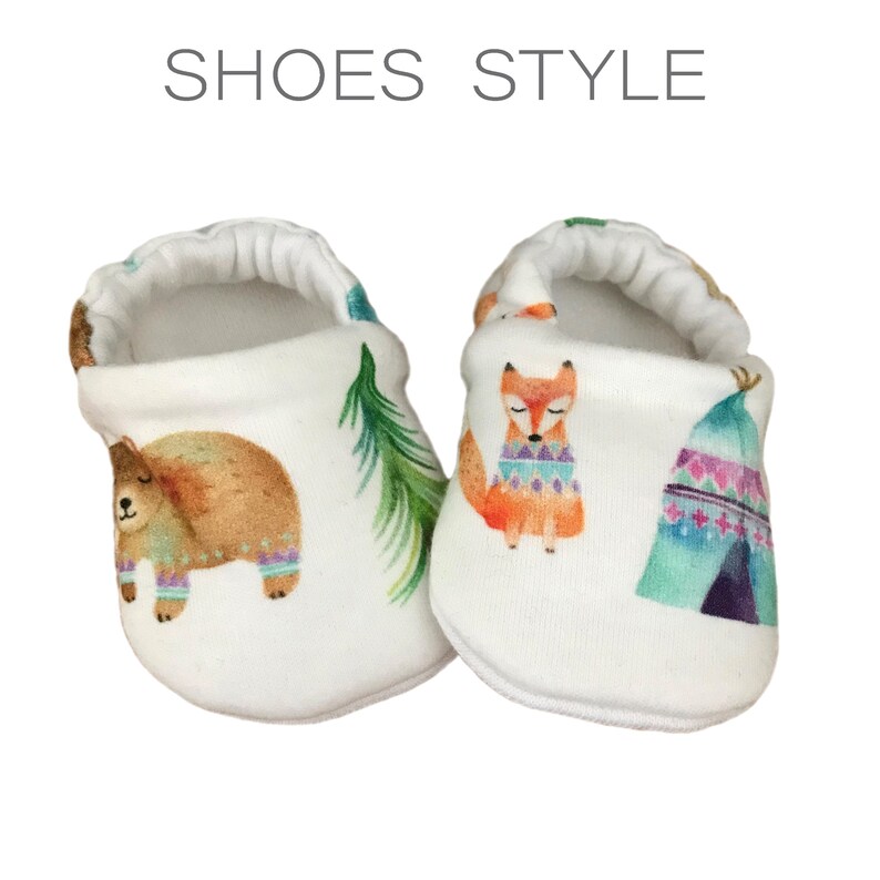 May include: A pair of white baby shoes with a woodland animal print. The shoes feature a bear, a fox, and a teepee with colorful geometric patterns.