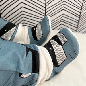 Op de afbeelding: Een paar babyschoentjes met een blauw en wit patroon van een wasbeer. De schoentjes zijn gemaakt van zacht materiaal en hebben een witte boord.