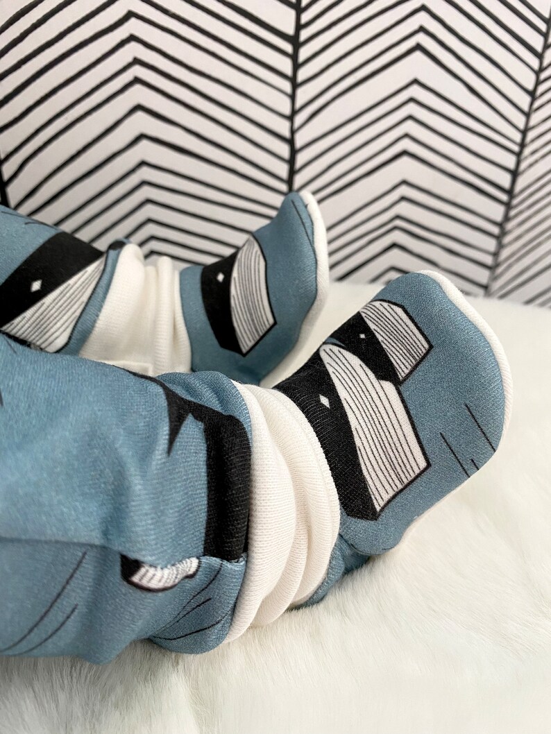 Op de afbeelding: Een paar babyschoentjes met een blauw en wit patroon van een wasbeer. De schoentjes zijn gemaakt van zacht materiaal en hebben een witte boord.