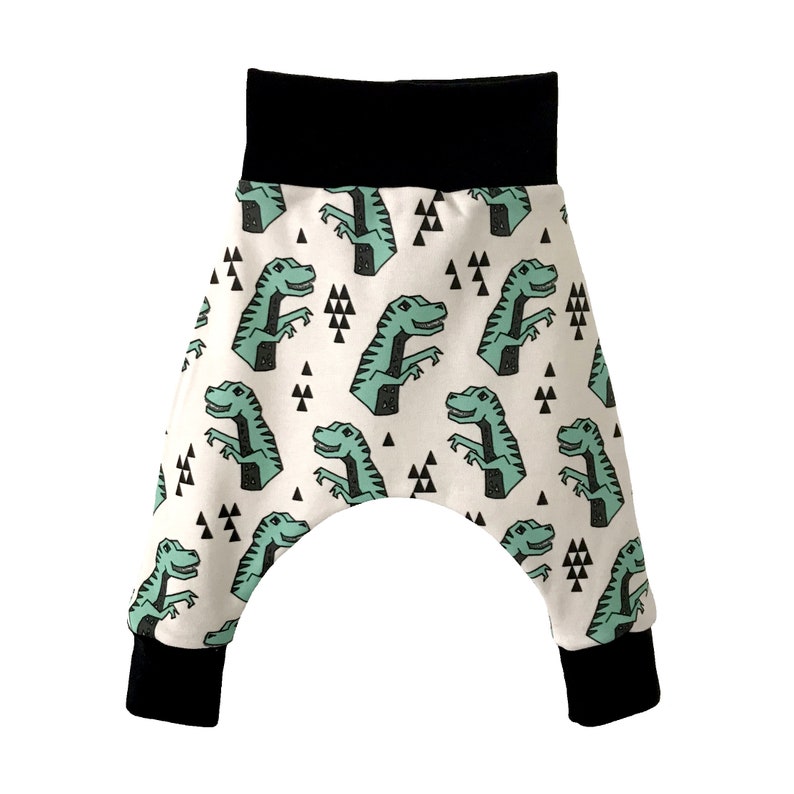May include: White baby pants with a black waistband and black cuffs. The pants have a green dinosaur print with black triangles.