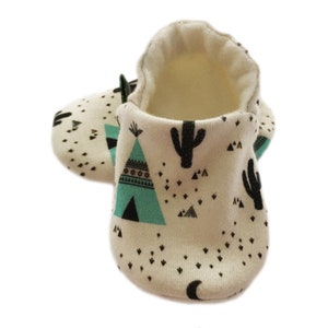 Könnte beinhalten: Ein Paar Babyschuhe mit weißem Hintergrund und einem Muster aus türkisfarbenen Tipis, schwarzen Kakteen und schwarzen Dreiecken. Die Schuhe haben ein weiches, flauschiges Futter.