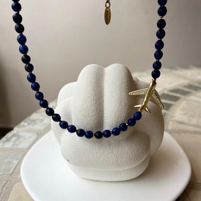May include: A blue lapis lazuli beaded necklace with a gold airplane charm. The necklace is on a white surface.