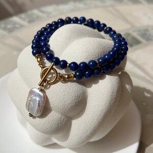 May include: A double-strand necklace made with dark blue lapis lazuli beads and a gold toggle clasp. A large, white baroque pearl hangs from the clasp.