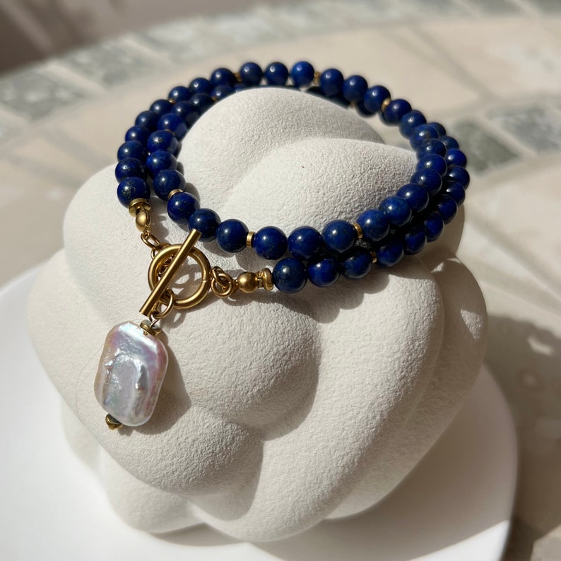 May include: A double-strand necklace made with dark blue lapis lazuli beads and a gold toggle clasp. A large, white baroque pearl hangs from the clasp.