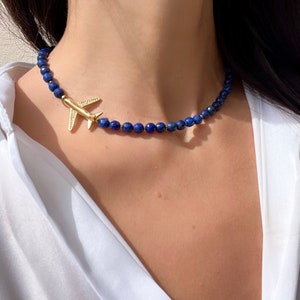 May include: A gold airplane charm hangs from a blue lapis lazuli beaded necklace. The necklace is worn by a person with dark hair.