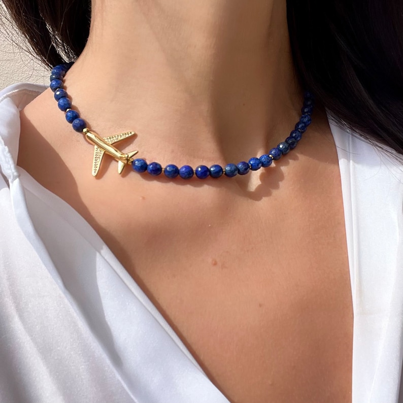 May include: A gold airplane charm hangs from a blue lapis lazuli beaded necklace. The necklace is worn by a person with dark hair.