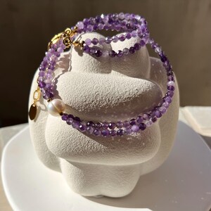 以下が含まれることがあります： 小さな紫色のファセットアメジストビーズを使用した、多層ブレスレット。1つの白い真珠と金色の留め金がコントラストを生み出しています。ブレスレットは白い彫刻された形の上に展示されており、日光が影を落としています。