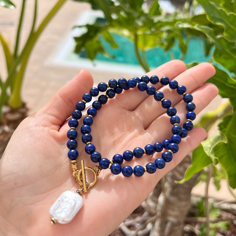 May include: A gold chain necklace with a large, white, rectangular pearl pendant. The chain is made of small, dark blue beads.