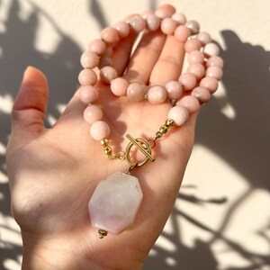 May include: A pink gemstone necklace with a large, faceted, pink gemstone pendant. The necklace has a gold toggle clasp.