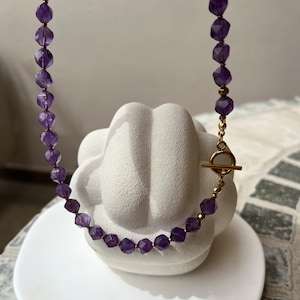 May include: A purple amethyst gemstone necklace with a gold toggle clasp.