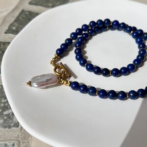 May include: A necklace made of dark blue lapis lazuli beads with a gold toggle clasp and a white pearl pendant.