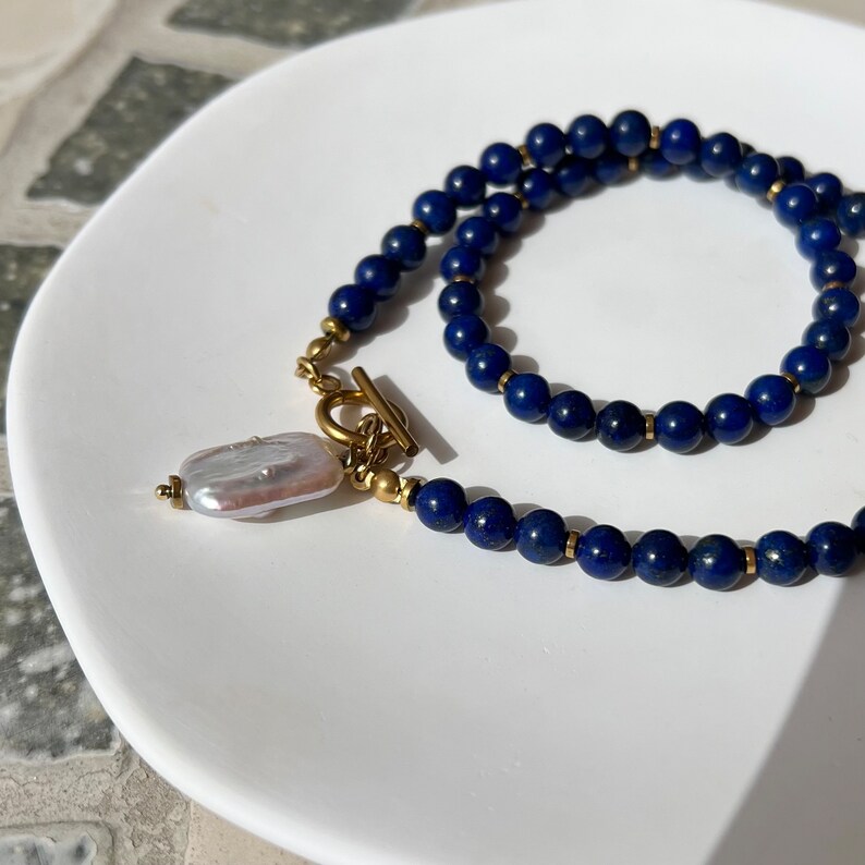 May include: A necklace made of dark blue lapis lazuli beads with a gold toggle clasp and a white pearl pendant.