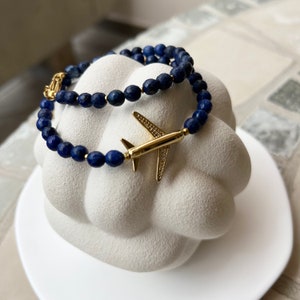 May include: Two blue lapis lazuli beaded bracelets with a gold airplane charm. The bracelets are stacked on top of each other and are displayed on a white surface.