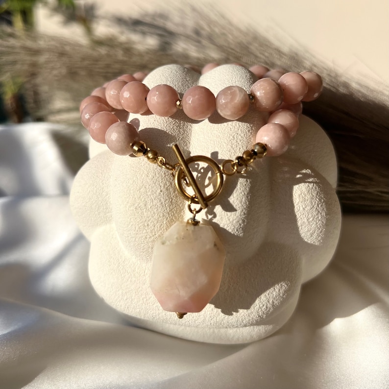 May include: A pink gemstone necklace with a large pink gemstone pendant. The necklace is made of gold-toned metal and has a toggle clasp.