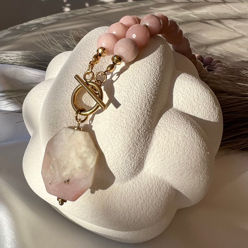 May include: A gold chain necklace with a large pink and white gemstone pendant. The necklace is made of small pink and white beads and has a gold toggle clasp.
