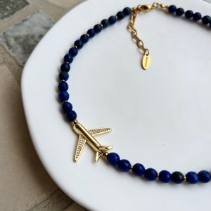 May include: A gold airplane charm with clear rhinestones hangs from a necklace made of dark blue lapis lazuli beads.
