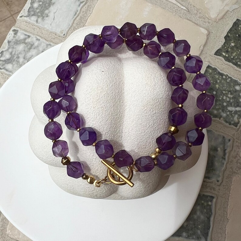 May include: A double-strand necklace made of amethyst gemstones and gold-toned metal. The necklace features a toggle clasp.