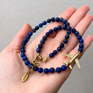 May include: A gold necklace and bracelet set featuring blue lapis lazuli beads and a gold airplane charm with clear rhinestones. The necklace has a gold clasp with the word "Momogram" engraved on it.
