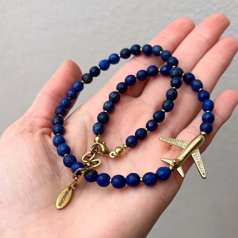 May include: A gold necklace and bracelet set featuring blue lapis lazuli beads and a gold airplane charm with clear rhinestones. The necklace has a gold clasp with the word "Momogram" engraved on it.