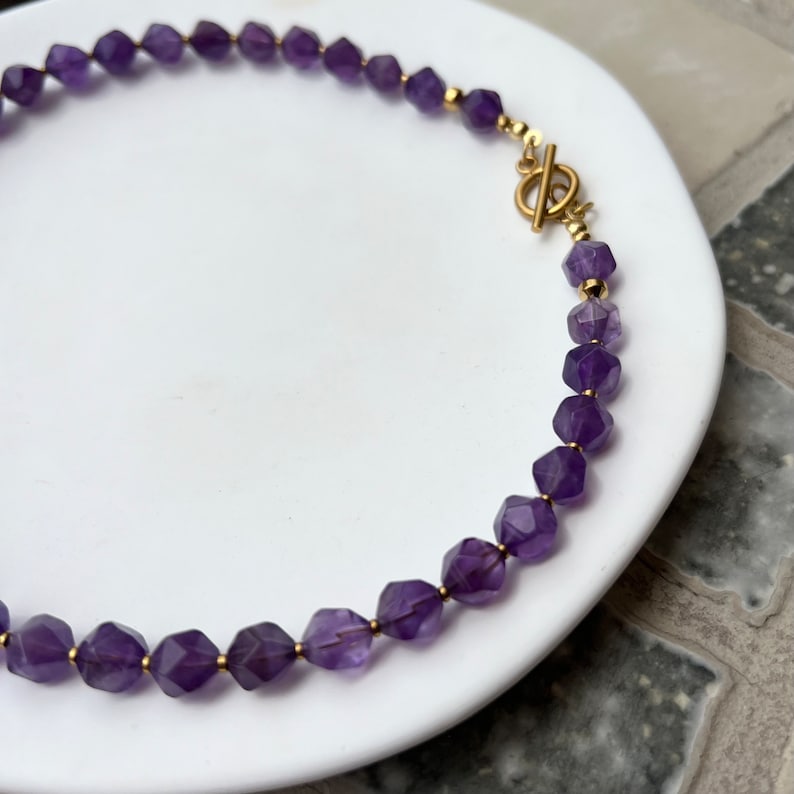 May include: A purple amethyst gemstone necklace with a gold clasp. The necklace is made of faceted amethyst beads.