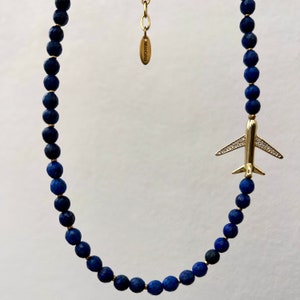 May include: A gold airplane charm hangs from a necklace made of dark blue lapis lazuli beads. The necklace has a gold clasp and a small tag that reads "Monogram".