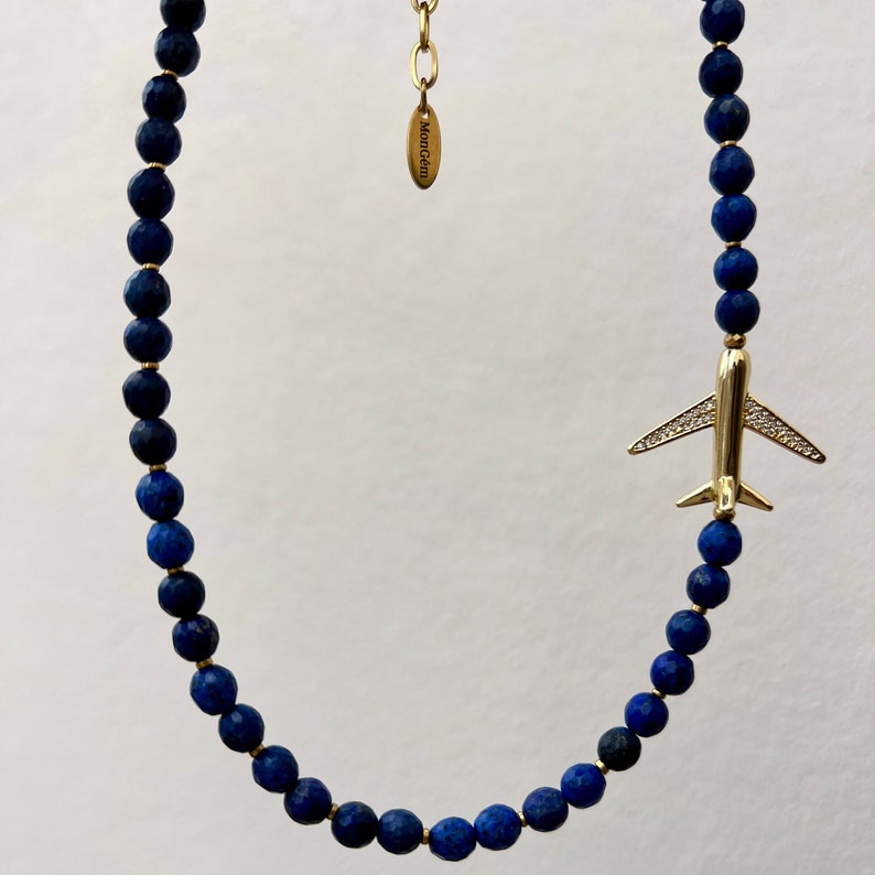 May include: A gold airplane charm hangs from a necklace made of dark blue lapis lazuli beads. The necklace has a gold clasp and a small tag that reads "Monogram".