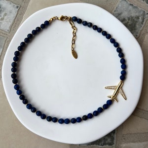 May include: A gold airplane charm hangs from a necklace made of dark blue lapis lazuli beads.