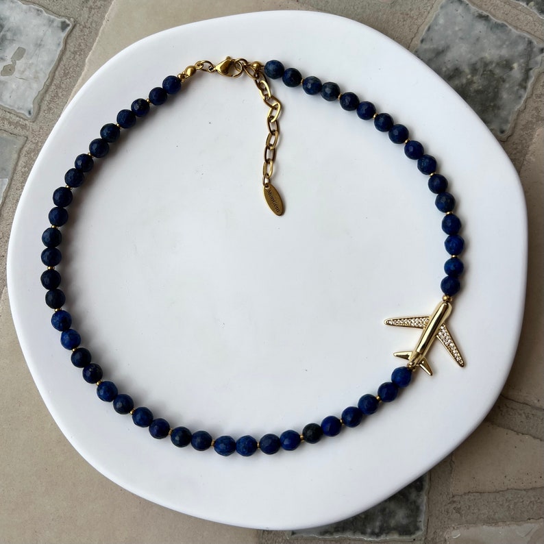 May include: A gold airplane charm hangs from a necklace made of dark blue lapis lazuli beads.