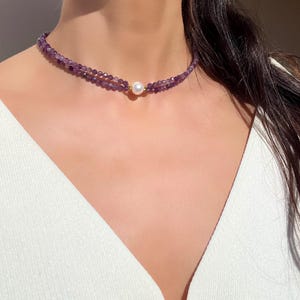 以下が含まれることがあります： 小さな紫のアメジストのビーズが特徴の繊細なチョーカーネックレス。 ネックレスは二重のストランドデザインで、中心に丸い白い真珠が1つあります。 金色のアクセントが見えます。