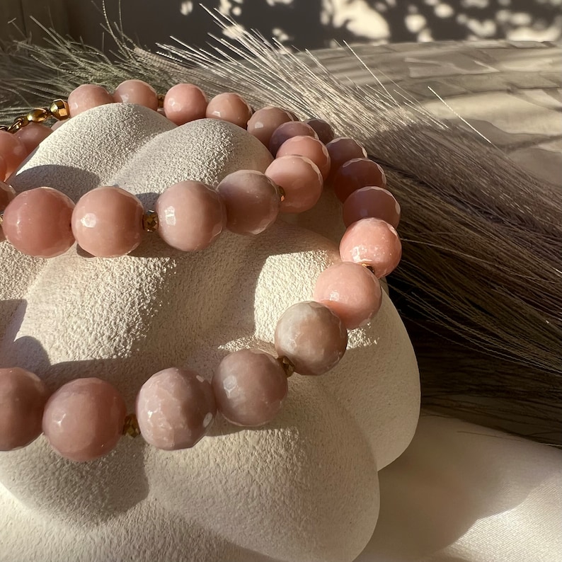 May include: A pink gemstone bracelet with gold accents. The bracelet is made of round, faceted beads and is displayed on a white, textured surface.