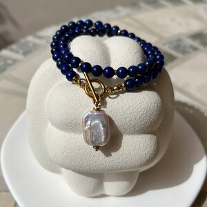May include: A double-strand necklace made with dark blue lapis lazuli beads and a gold toggle clasp. A large, white baroque pearl hangs from the clasp.