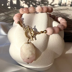 May include: A pink gemstone necklace with a large, faceted, pink and white gemstone pendant. The necklace has a gold toggle clasp.