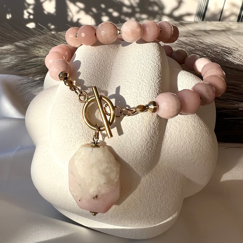 May include: A pink gemstone necklace with a large, faceted, pink and white gemstone pendant. The necklace has a gold toggle clasp.